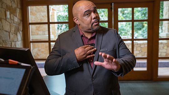 Businessman in a dark suit gesturing while speaking at a podium in a bright room with large windows behind him.