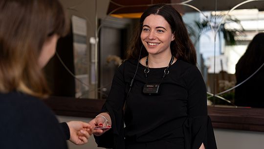 Smiling staff member in black with a name badge hands a card to a visitor at a reception desk.