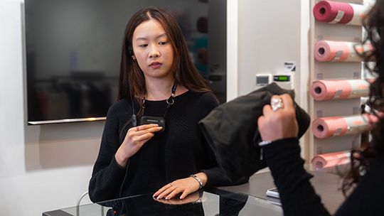 Customer at a store checkout counter glances at her smartphone while a cashier hands over a black bag