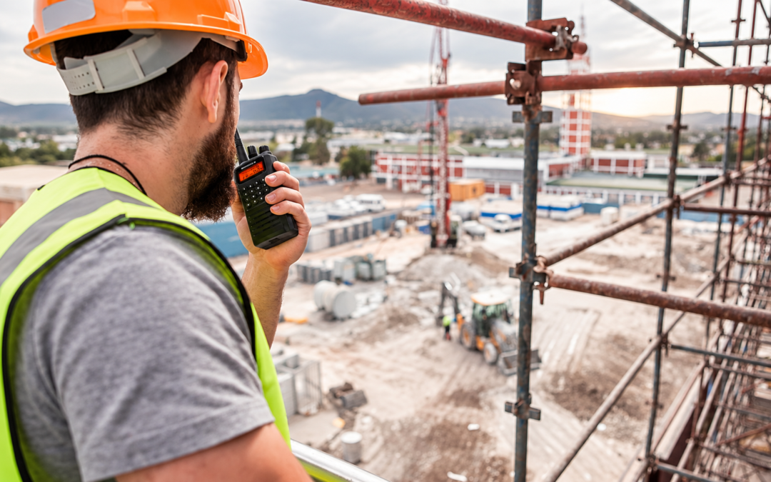 Construction worker in a safety vest and orange hard hat talks on a handheld radio on a scaffolded construction site.